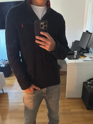 Mörkblå kabelstickad tröja Polo Ralph Lauren - Mörkblå kabelstickad tröja från Polo Ralph Lauren med halv dragkedja och röd logga på bröstet. Tröjan har ribbstickad krage med röd och marinblå randig insida samt långa ärmar. Perfekt för en stilren och klassisk look.