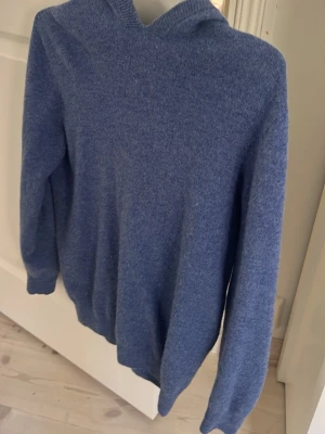 Blå hoodie i mjukt stickat material - En stilren blå hoodie i mjukt stickat material med lång ärm och ribbade muddar. Perfekt för dig som gillar en enkel och avslappnad look. Huvan ger extra mysfaktor och passar till många olika outfits.