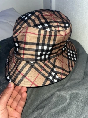 Burberry hat o kenzo tröja - Snygg bucket hat i beige med klassiskt rutigt mönster i svart, vitt och rött. Hatten har bred brätte och är tillverkad i ett slätt tyg. Perfekt accessoar för att lyfta din outfit.