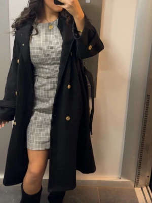 Grå rutig kortklänning - Säljer en grå och vit rutig kortklänning med tight passform och korta ärmar. Klänningen har en klassisk, stilren look och är tillverkad i ett mjukt bomullsliknande material. Perfekt för dig som gillar trendiga och bekväma outfits. Använd en gång