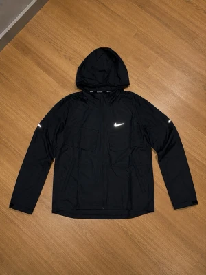Svart Nike träningsset med huva - Säljer ett svart träningsset från Nike med matchande jacka och byxor. Jackan har huva, dragkedja och reflexdetaljer samt två bröstfickor. Byxorna har resår i midjan och vid benslut samt en vit Nike-logga. Perfekt för löpning eller gym.