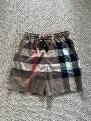 Beige-rutiga shorts med snörning - Säljer ett par beige shorts med klassiskt rutigt mönster i svart, vitt, blått och rött. Shortsen har elastisk midja med svart snörning och sidofickor samt en bakficka. Perfekta för sommaren och riktigt snygga till en enkel t-shirt.