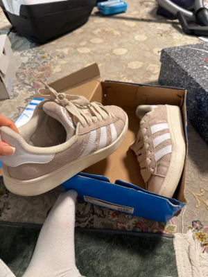 Beige Adidas Campus sneakers - Snygga beige Adidas Campus sneakers med klassiska vita ränder på sidan. Skorna har en låg profil och är gjorda i mocka med vita detaljer i läder. Perfekta för en avslappnad och trendig stil. Priset går att diskutera 