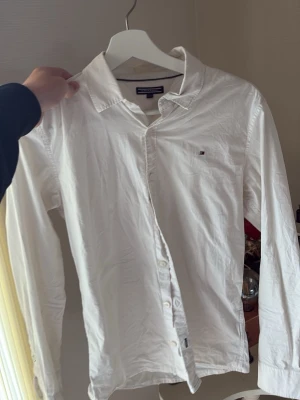 Vit skjorta från Tommy Hilfiger - Klassisk vit skjorta från Tommy Hilfiger med lång ärm och knappar framtill. Skjortan har en liten broderad logga på bröstet och är tillverkad i bomull. Perfekt för en clean och stilren look.