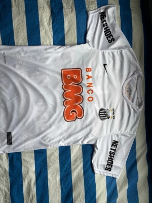 Vit Santos FC Neymar matchtröja - Säljer en vit Santos FC matchtröja från Nike med Neymar Jr och nummer 11 på ryggen. Tröjan har tryckta sponsorer som BMG och Netshoes, klubbmärke på bröstet och rund hals. Perfekt för dig som älskar brasiliansk fotboll och vill ha en ikonisk supportertröja. Endast använd 1 gång men säljes pga för liten för mig. 