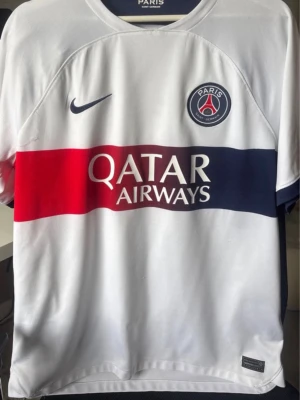 PSG vit matchtröja med röd/blå rand - Säljer en vit PSG matchtröja från Nike med bred röd och marinblå rand över bröstet. PSG-logga och Nike-swoosh på bröstet samt Qatar Airways-tryck. Tillverkad i lätt och ventilerande material, perfekt för fotboll eller träning.