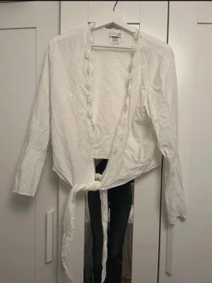 Vit topp med knytband - Vit omlottblus med långa ärmar och knytband i midjan. Blusen har volangdetaljer längs kanten och är gjord i ett tunt, lätt tyg. Perfekt för en stilren och trendig look.