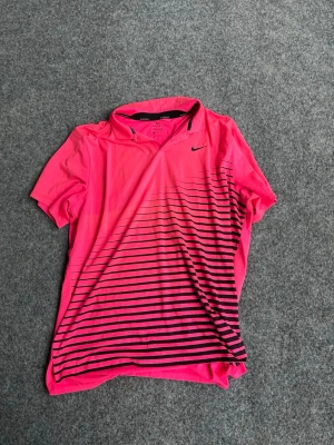 Rosa Nike t-shirt med svarta ränder - Snygg rosa t-shirt från Nike med svarta horisontella ränder nertill på framsidan. T-shirten har korta ärmar, klassisk krage och är tillverkad i ett lätt och sportigt material som passar perfekt för träning eller tennis.