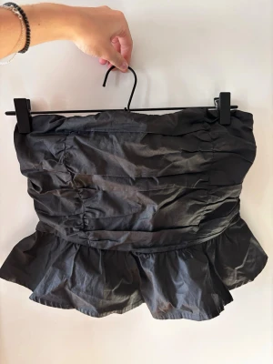 Svart axelbandslös volangtopp - Säljer en svart axelbandslös topp med volangkant nedtill. Toppen har en croppad passform. Snygga rynkade detaljer framtill och baktill ger en cool och trendig look. Passar toppen till fest, t.ex. nyår! Tyvärr helt oänvänd.