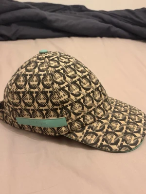 Ferregamo Keps - Säljer en snygg och unik modell  ferregamo keps med beige och svart grafiskt mönster. Kepsen har böjd skärm och detaljer i ljusgrönt, bland annat på justerbar rem bak och på toppen. 
