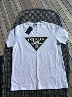 Vit t-shir Prada - Säljer en vit t-shirt från Prada med det ikoniska svarta triangulära logotrycket på bröstet. Klassisk passform och rund halsringning. Perfekt för dig som vill ha en clean och stilren look med exklusiv känsla.