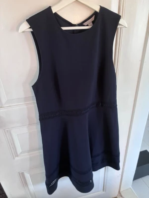 Mörkblå ärmlös kortklänning från H&M - Snygg mörkblå ärmlös klänning med rund halsringning och dekorativa band i midjan och vid fållen. Klänningen har en klassisk, enkel siluett och stängs med dragkedja bak. Perfekt för dig som gillar stilrena plagg.