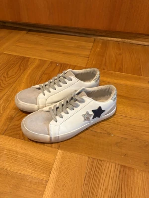 Vita sneakers med stjärndetaljer - Snygga vita sneakers med gråa skosnören och tåparti i mockaliknande material. På sidan finns två stjärnor, en i silverglitter och en i svart. Skon har lågt skaft och silverdetaljer vid hälen. Perfekta för en trendig och avslappnad look.