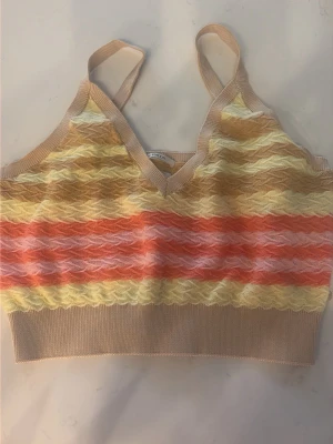 Stickad randig croppad linnetopp - Säljer en croppad stickad linnetopp med smala axelband och v-ringning. Toppen har vågiga ränder i beige, gul, orange och ljusrosa. Perfekt för sommarens alla tillfällen och ger en färgglad touch till din outfit.