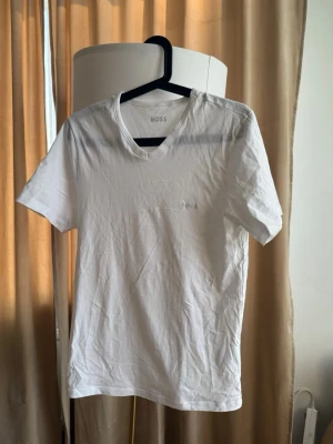 Vit t-shirt från BOSS - Klassisk vit t-shirt från BOSS med diskret logga på bröstet. T-shirten har korta ärmar och v-ringad hals. Tillverkad i mjuk bomull för en skön känsla och enkel stil som funkar till allt.
