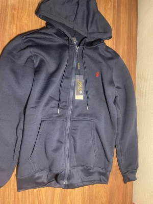 Polo Ralph lauren hoodie  - Snygg mörkblå hoodie med dragkedja och huva. Har två stora fickor framtill och en liten röd logga broderad på bröstet. Tillverkad i 100% polyester, perfekt för en chill och avslappnad stil. Dragsko i huvan för justerbar passform. Skriv vid minsta funderingar. 
