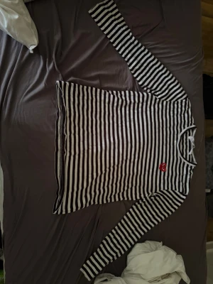 commes des garcons tröja  - Säljer en svartvit randig långärmad topp med rund hals och en röd hjärtdetalj på bröstet. Perfekt för dig som gillar klassiska mönster med en twist. Passar till både jeans och kjol för en cool vardagslook.