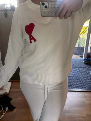 Ami tröja med rött hjärta - Vit stickad tröja med ett stort rött hjärta och bokstaven 'A' broderat på bröstet. Tröjan har rund halsringning, långa ärmar och breda ribbade muddar vid ärmslut och nederkant. Perfekt för dig som gillar enkel men snygg design.