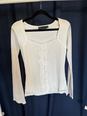 Vit långärmad topp med spets KappAhl - Säljer en vit långärmad topp från KappAhl med fina spetsdetaljer längs ringningen och framtill. Toppen har en feminin och enkel design med lätt stretchigt material och rundad hals. Perfekt att styla med jeans eller kjol.