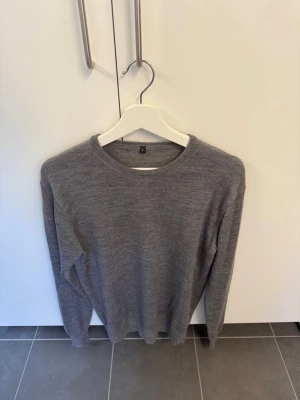 Grå stickad tröja i ull, storlek L i  - En enkel och stilren grå stickad tröja med rund halsringning. Tröjan har långa ärmar och ribbade muddar vid hals, ärmslut och nederkant. Perfekt basplagg för lager på lager och passar till det mesta.