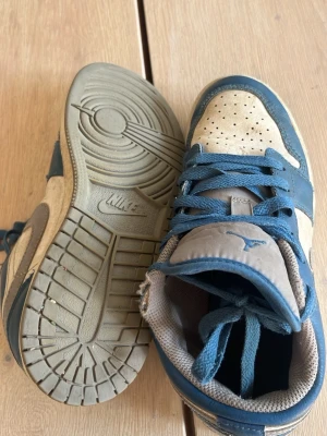 Nike blå och beige sneakers - Snygga sneakers från Nike i blått och beige med klassisk låg siluett. Skorna har perforerad tåbox, blå snörning och detaljer i läder och textil. Yttersulan är i gummi med tydlig Nike-logga undertill. Perfekta för en avslappnad streetwear-look.