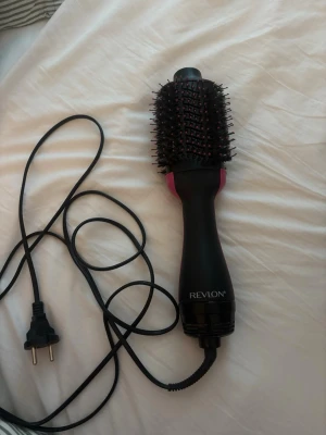 Revlon svart varmluftsborste - En svart varmluftsborste från Revlon med rund borste och täta borststrån. Handtaget är ergonomiskt och har rosa detaljer. Perfekt för att styla och torka håret samtidigt. Elektrisk med lång sladd och europeisk kontakt.
