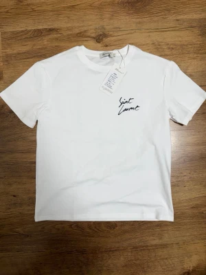 Vit Saint Laurent t-shirt - Snygg vit t-shirt från Saint Laurent med svart logotyp på bröstet. Klassisk rund hals och korta ärmar. Tillverkad i mjukt material som känns skönt mot huden. Perfekt för dig som gillar stilrena och trendiga plagg.Står storlek S på tröjan men passar Xs