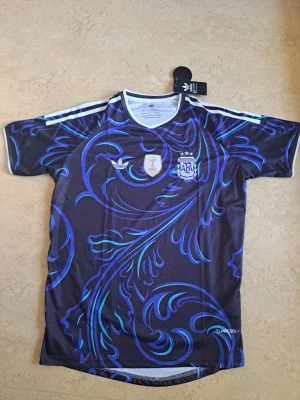 Argentina fotbollströja - Snygg Argentina fotbollströja från Adidas i mörkblått med blått och turkost mönster. Tröjan har vita ränder på axlarna, broderade märken och Climalite-material som andas. Perfekt för träning eller match. Helt ny med prislappar.