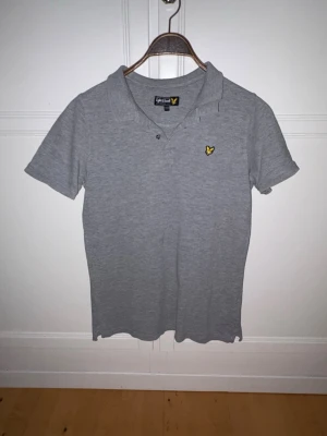 Grå pikétröja Lyle & Scott - Säljer en stilren grå pikétröja från Lyle & Scott med klassisk krage och korta ärmar. Tröjan har en gul logga på bröstet och knäppning vid halsen. Perfekt för en avslappnad och snygg look.