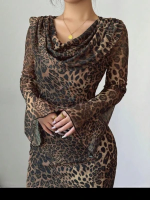 Leopardmönstrad långklänning - Snygg långklänning med leopardmönster och utsvängda ärmar. Klänningen har en draperad urringning och är figurnära med transparenta ärmar. Perfekt för dig som vill sticka ut med en trendig look.