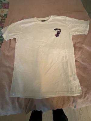 Vit Stüssy t-shirt med tärningsmotiv - Vit t-shirt från Stüssy med tryck av två lila tärningar och Stüssy-loggan på bröstet. Klassisk rund hals och korta ärmar. Perfekt för dig som gillar streetwear och vill ha en enkel men snygg tee med cool detalj.