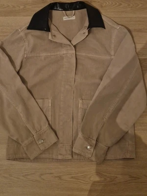 Beige Jeans jacka - Jacka från Gina Tricot. Perfekt nu för sommaren/ hösten. Stilren beige jacka med kontrasterande svart krage och stora framfickor. Jackan har långa ärmar, knappar vid ärmsluten och är tillverkad i ett kraftigare bomullstyg. Perfekt för dig som gillar minimalistisk och clean stil.