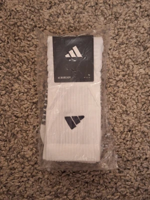 Vita Adidas strumpor AEROREADY - Fräscha vita strumpor från Adidas med svart logga på skaftet. Storlek: 43-45. Modellen är AEROREADY som andas och är perfekta för dig som vill ha bekväma och sportiga strumpor. Ribbad struktur upptill för extra komfort. Skick: 10/10