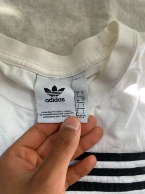 Vit Adidas t-shirt  - Vit t-shirt från Adidas med klassiska svarta ränder över bröstet och Adidas-logga framtill. T-shirten har rund halsringning och är tillverkad i mjuk bomull. Perfekt för en sportig och stilren look.
