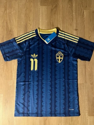 Elanga Sverige vm S - Säljer en blå fotbollströja för Sverige med Elanga och nummer 11 på ryggen. Tröjan har gula detaljer, korta ärmar och ett subtilt mönster. Adidas-logga och svensk landslagssköld på bröstet. Perfekt för fotbollsfans!