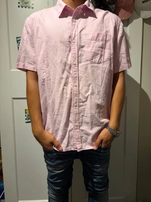 Ljusrosa kortärmad skjorta - Säljer en ljusrosa skjorta med korta ärmar och klassisk krage. Skjortan har knappar framtill och en bröstficka. Perfekt för en avslappnad och stilren look.