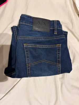 Mörkblå jeans från Armani Exchange - Snygga mörkblå jeans från Armani Exchange med klassisk femficksdesign och kontrastsömmar i gult. Jeansen har en rak passform och är tillverkade i ett mjukt denimtyg. Perfekta för en stilren och avslappnad look.