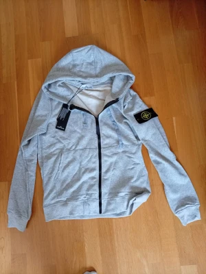 Grå stone island hoodie - Snygg grå hoodie med dragkedja framtill och huva. Tröjan har en svart patch med gul och grön brodyr på vänster ärm, samt fickor på sidorna. Tillverkad i mjukt sweatshirtmaterial som är perfekt för chill dagar. Passar dig som gillar streetwear och enkel stil.
