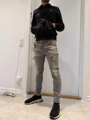Grå slitna Dsquared 2 jeans herr. Dsq2 storlek 42  - Säljer ett par grå Dsquared 2 jeans med slitningar och detaljerade revor på benen. Jeansen har klassisk femficksdesign och dragkedjegylf. Snygg passform som sitter tight hela vägen ner. Perfekta för en trendig streetstil.