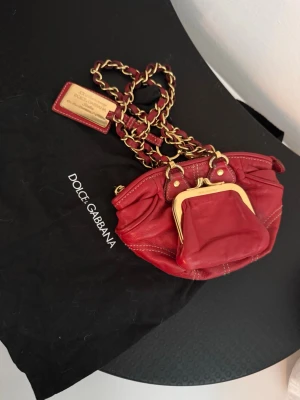 Röd handväska från Dolce & Gabbana - Snygg röd handväska i äkta läder från Dolce & Gabbana med guldfärgade kedjedetaljer och en liten matchande myntbörs framtill. Väskan har leopardmönstrat innerfoder och exklusiva metalldetaljer. Perfekt för dig som vill sticka ut med en lyxig accessoar.