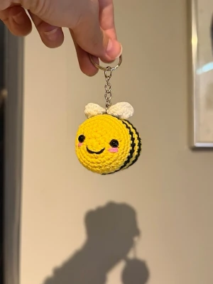 Handvirkad bi nyckelring  - Supersöt handvirkad nyckelring i form av ett bi. Biet är gult med svarta ränder, vita vingar och har ett gulligt ansikte med broderade ögon och rosa kinder. Perfekt att hänga på väskan eller nycklarna för en färgglad touch.