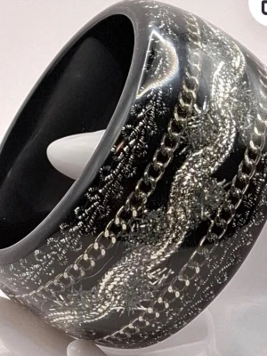 Brett svart armband med silverdetaljer - Snyggt brett armband i svart plast med glansig finish och inlagda silverfärgade kedjor och mönster. Armbandet har en cool, chunky look och passar perfekt för dig som vill sticka ut med accessoarerna. Materialet är plast med metalliska detaljer.