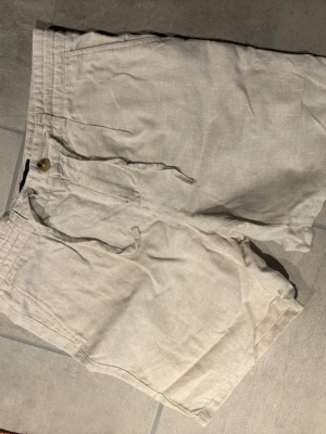 Beiga linneshorts med knytband - Snygga beiga shorts i linne med knytband i midjan och knappstängning. Klassisk modell med fickor fram och bak, perfekta för varma dagar.
