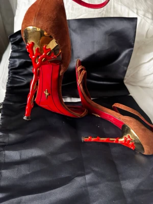 Cesare Paciotti! Bruna pumps med röda klackdetaljer - Unika pumps i brun mocka med öppen tå och spektakulära röda klackar som liknar korallgrenar. Guldfärgade detaljer vid hälen och röd insida ger en lyxig känsla. Perfekta för dig som vill sticka ut med ett par riktigt statement-heels.