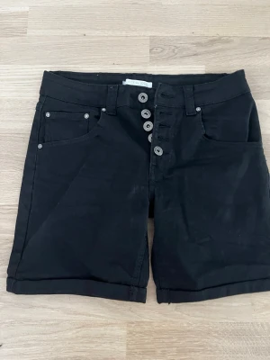 Svarta shorts från LACE du JOUR - Snygga svarta shorts från LACE du JOUR med uppvikta benslut och fem knappar framtill. Klassisk femficksmodell och midwaist passform. Perfekta för en avslappnad och trendig look. Strl 36