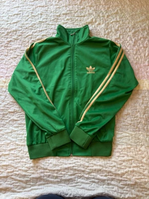 Grön Adidas track jacket - Snygg grön Adidas track jacket med guldiga ränder längs ärmarna och dragkedja framtill. Klassisk Adidas-logga på bröstet och stort tryck på ryggen. Ribbstickade muddar vid ärmslut och nederkant. Perfekt för en sportig och retro look.