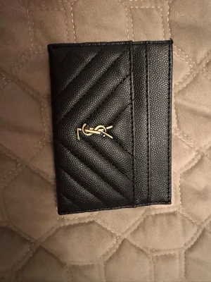 Svart quiltad kortplånbok YSL - Snygg svart kortplånbok från YSL i quiltat läder med guldfärgad logga på framsidan. Plånboken har flera kortfack och en stilren, modern design som passar perfekt i fickan eller väskan.