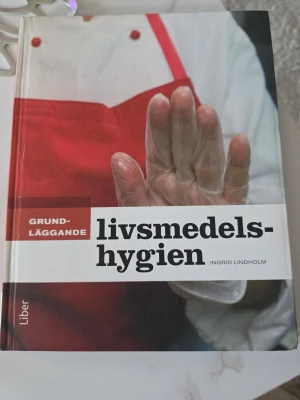Grundläggande livsmedelshygien - Boken 'Grundläggande livsmedelshygien' av Ingrid Lindholm ger en tydlig genomgång av hygienregler, mikroorganismer och livsmedelssäkerhet. Omslaget är rött och vitt med en person i plastförkläde och plasthandske. Utgiven av Liber.