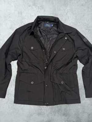 Svart vindjacka från Polo Ralph Lauren - Snygg svart vindjacka från Polo Ralph Lauren med fyra framfickor, dragkedja och knappar. Jackan har hög krage, justerbar midja och diskret logga på ärmen. Tillverkad i ett lätt, vattenavvisande material som passar perfekt för blåsiga dagar. Bredd - 50cm, Längd - 74cm, Ärm - 64cm. Passar mig bra som är 181cm lång. Byten av andra klädesplagg, skor, prylar osv kan vara intressant så bara att höra av sig vid fundering! Mvh 