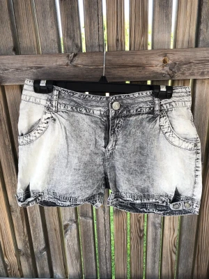 Grå tvättade jeansshorts - Säljer ett par grå tvättade jeansshorts med råa kanter och knappdetaljer vid bensluten. Shortsen har fem fickor, bälteshällor och en klassisk knappgylf. Perfekta för sommaren och ger en avslappnad streetstyle-vibe.